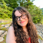 Person with long hair and glasses smiling outdoors.
