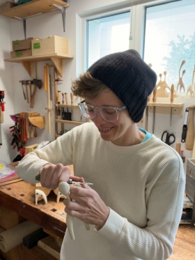 Headshot of Katie Hudnall. Person in black knitted cap and glasses smiling while sculpting a small wooden piece, indoors.