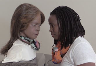 Image courtesy of the artist. Stephanie Dinkins, “Conversations with Bina48”, ongoing video project, 2014-present. Profile of person in white shirt and orange scarf staring directly at head/bust of robot figure with sculpted face, wig, white shirt, and mint green and pink scarf.