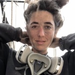 Image of Erica Compton. Person looking forward with hair pulled back wearing a black sweater and respirator dangling from neck. 