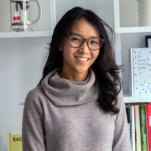 Person with long hair, glasses, and turtleneck sweater smiling indoors.