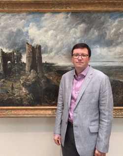Tim Barringer in a gray jacket and dust pink button-up shirt standing in front of a John Constable painting on display