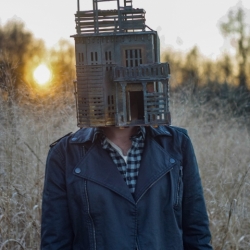 Person standing in field with sculpture of a house on their head, outdoors.