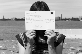 Black and White image of a person outdoors in front of a body of water with buildings on the horizon holding a postcard held up to their face.