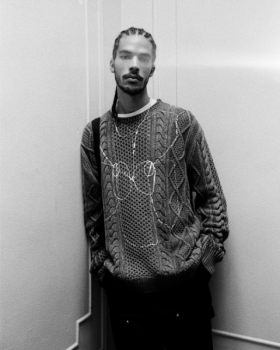 Headshot of Rahim Fortune. Person in braids and sweatshirts leaning against wall, indoors, black and white image.