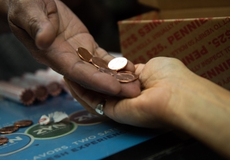 Vertical image of an outstretched hand with several pennies resting on a receiving hand over a counter laminated in a blue advertisement. An open brown box with diagonal red text 'PENNIES' and rolls of coins sealed in paper are visible in the background.