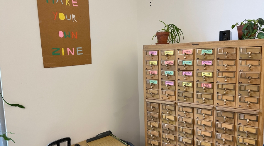 An Image of the Art Library's makerspace. There is a cabinet of drawers with materials for making zines. On a low table, there is a paper cutter and several hole punches. On the wall above the table there is a sign that says Make Your Zine.