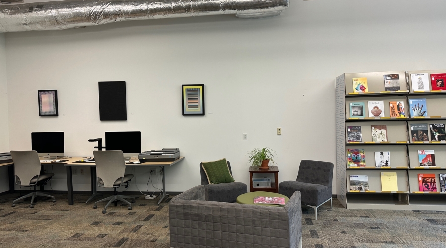 A view of the left or southern side of the Art Library. Centered is a couch and two chairs. To the left of the chairs, there are two desktop computers with scanners. To the right of the chairs, there is a series of periodical shelves.