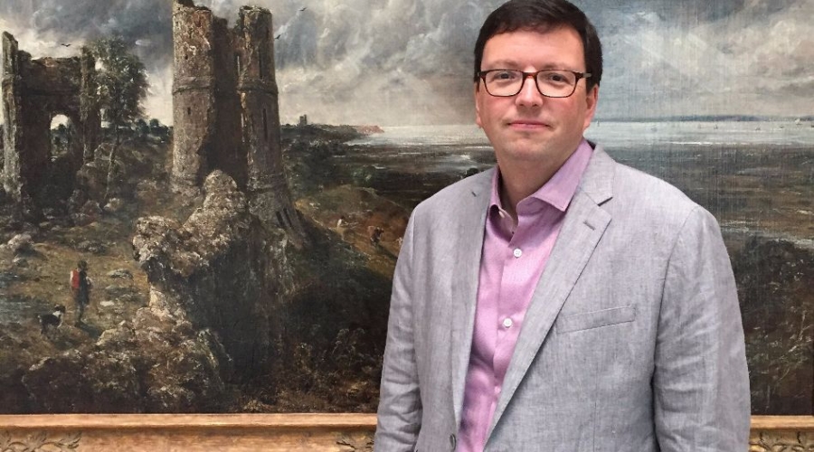 Tim Barringer in a gray jacket and dust pink button-up shirt standing in front of a John Constable painting on display