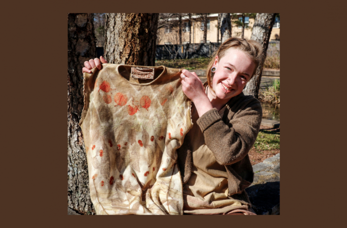 Individual outdoors smiling while holding up a brown dress with natural dyes