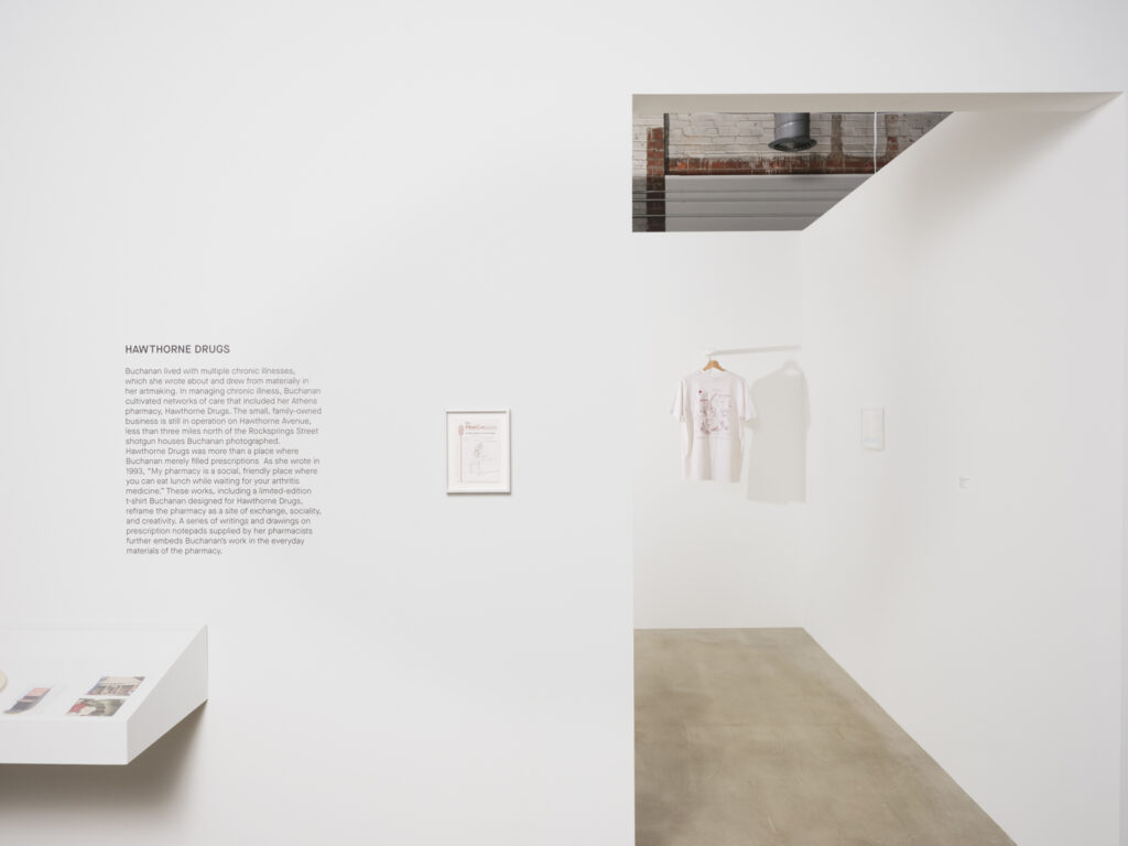 Installation view at the Athenaeum for “Beverly's Athens: Beverly Buchanan in Athens, GA”, Spring 2026. Entryway of white wall gallery installation with exhibition wall text in foreground and single suspended white t-shirt on a hanger in the background. Photo courtesy of Walker Bankson.