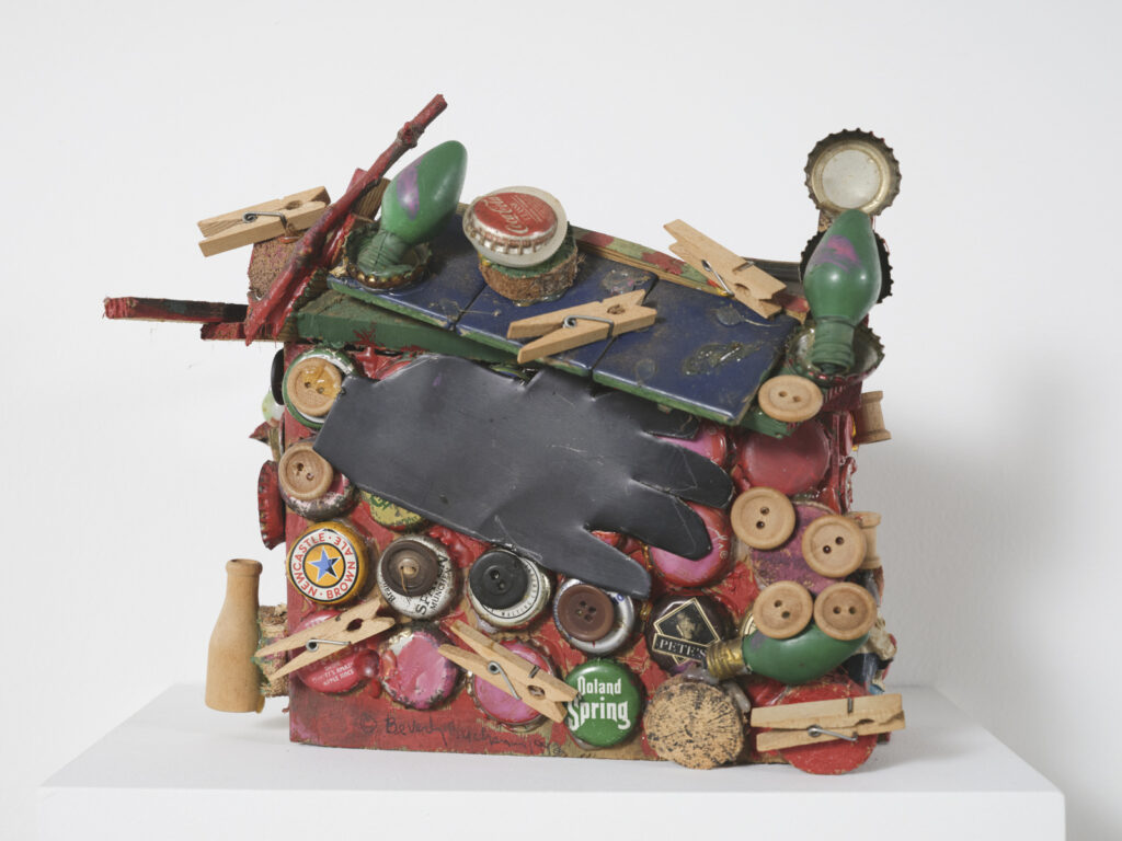Side view of assembled sculpture on white pedestal. Shack-like object is decorated with affixed bottle caps, buttons, a black glove, clothespins, and green plastic, among other objects on red painted side wall and forest green roof. Artwork by Beverly Buchanan on view at the Athenaeum for “Beverly's Athens: Beverly Buchanan in Athens, GA”, Spring 2026. Photo courtesy of Walker Bankson.