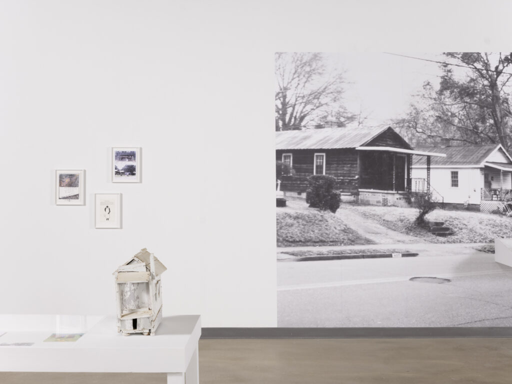 Installation view at the Athenaeum for “Beverly's Athens: Beverly Buchanan in Athens, GA”, Spring 2026. Small white shack sculpture on low white table on lower left. Wall behind the object includes a large print of a black and white photograph of houses along a street and small framed images and documents. Photo courtesy of Walker Bankson.