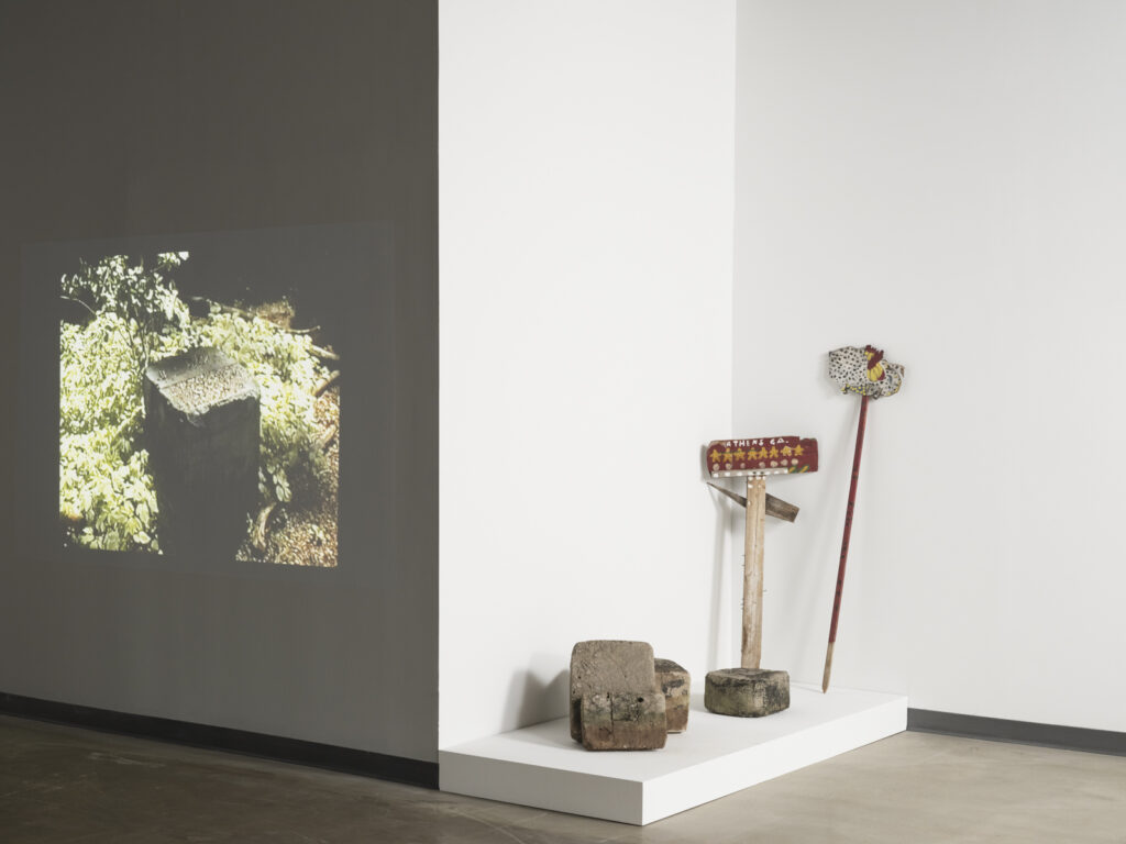 Installation view at the Athenaeum for “Beverly's Athens: Beverly Buchanan in Athens, GA”, Spring 2026. Small white shack sculpture on low white table on lower left. Wall behind the object includes a large print of a black and white photograph of houses along a street and small framed images and documents. Photo courtesy of Walker Bankson.